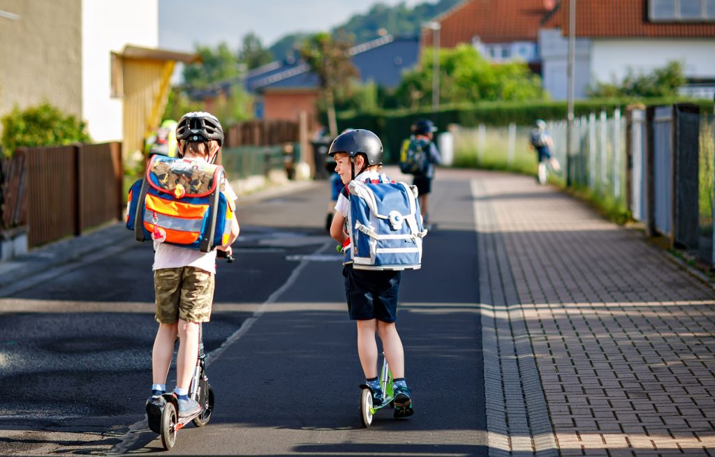 Two school kid boys in safety helmet riding with scooter in the city with backpack on sunny day. Happy children in colorful clothes biking on way to school. Safe way for kids outdoors to school