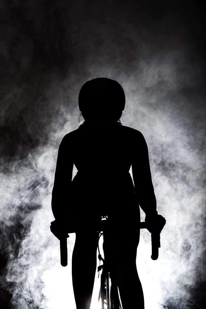 female roadbiker silhouette riding a bicycle against dark background with fog